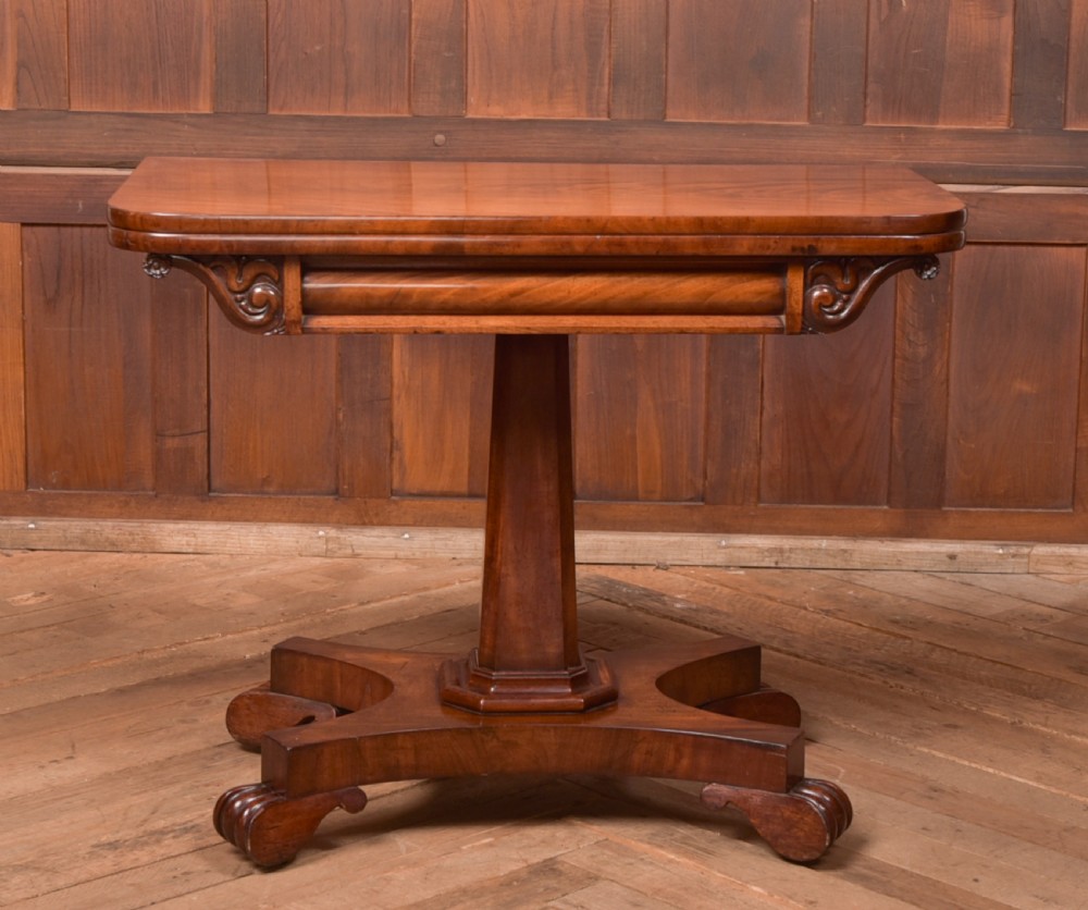 A Victorian mahogany card table featuring a rectangular fold-over top set upon a central tapered pedestal and a quadripartite plinth base with carved feet. View XVI