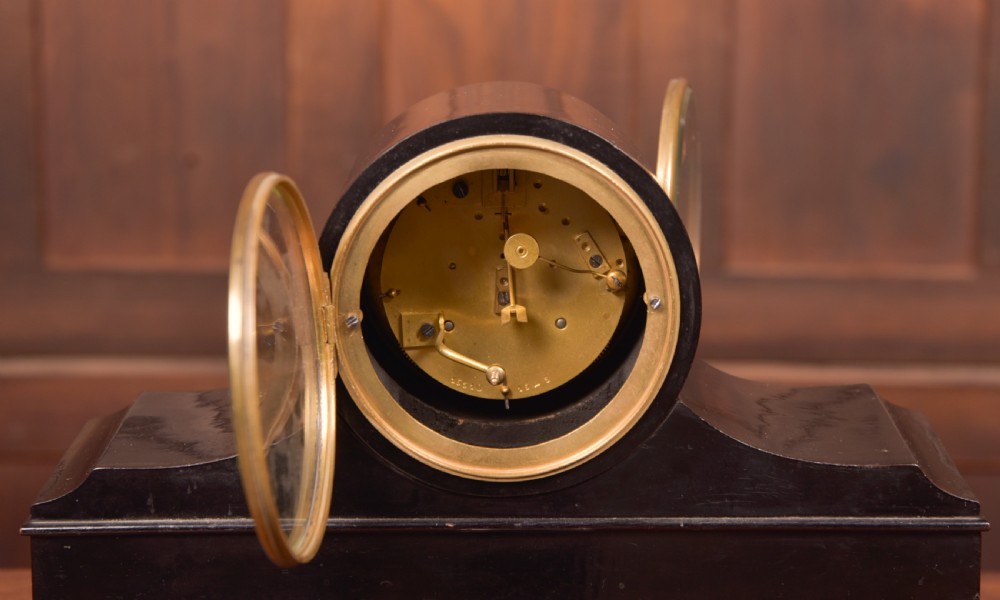 A Victorian black ebonised mantel clock featuring a circular brass-rimmed dial with Roman numerals set above a rectangular plinth base. View IX