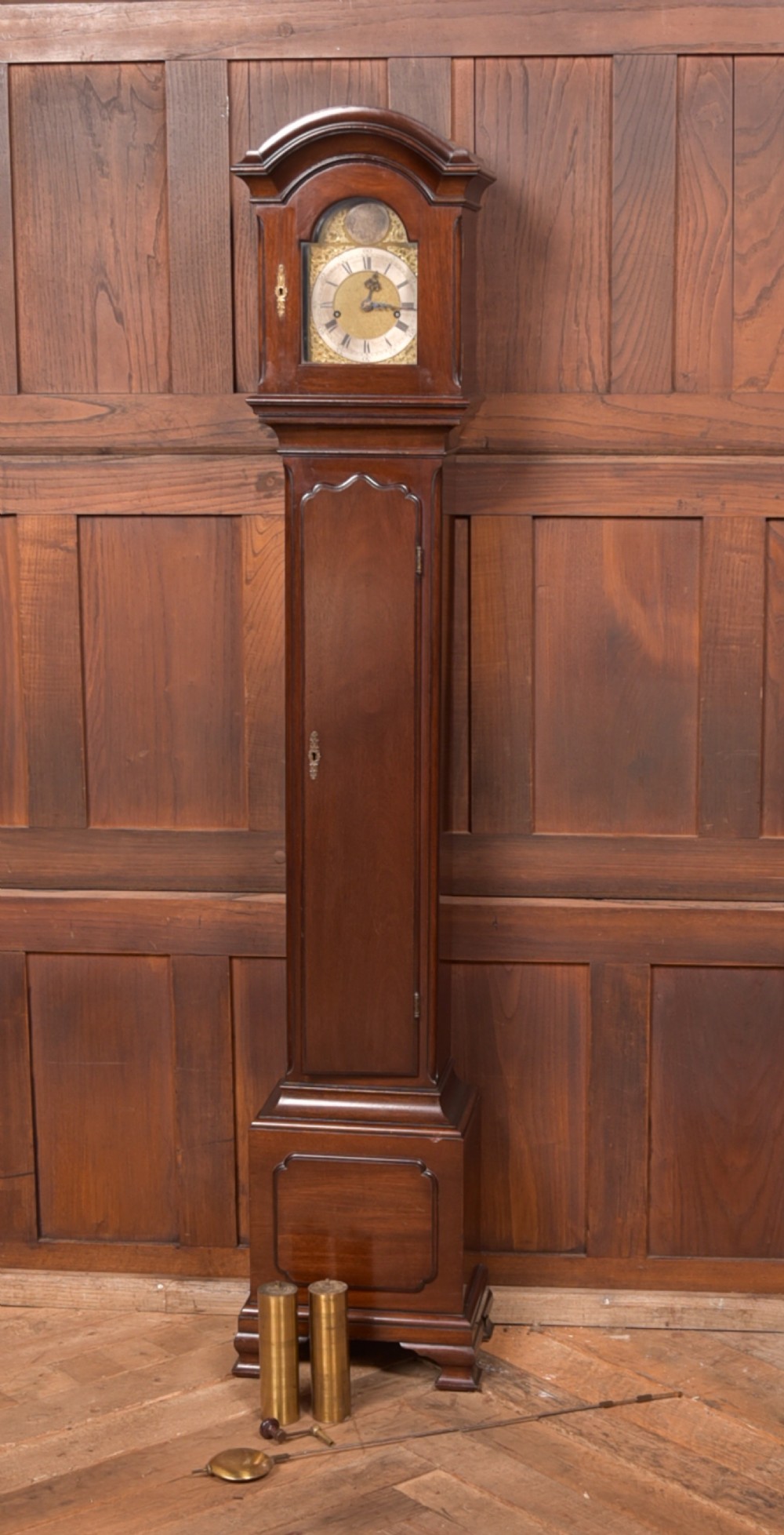A freestanding mahogany grandmother clock featuring an arched hood, brass dial with corner spandrels, a slender waist door, and brass weights placed in front.