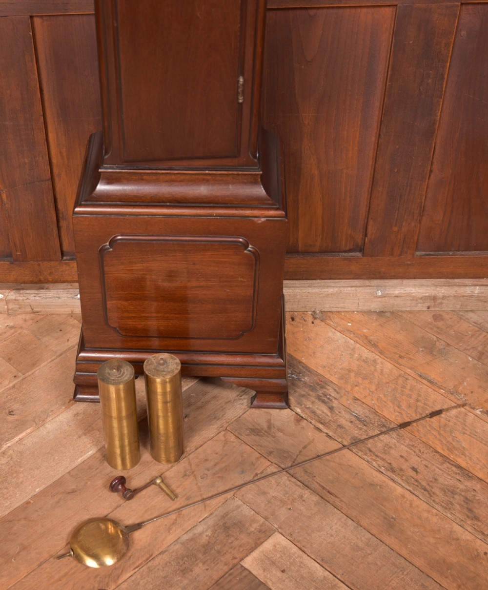 A freestanding mahogany grandmother clock featuring an arched hood, brass dial with corner spandrels, a slender waist door, and brass weights placed in front. View V
