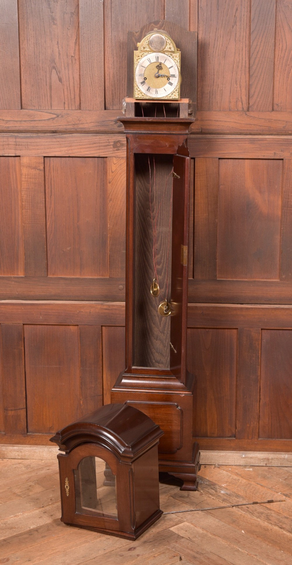 A freestanding mahogany grandmother clock featuring an arched hood, brass dial with corner spandrels, a slender waist door, and brass weights placed in front. View VII