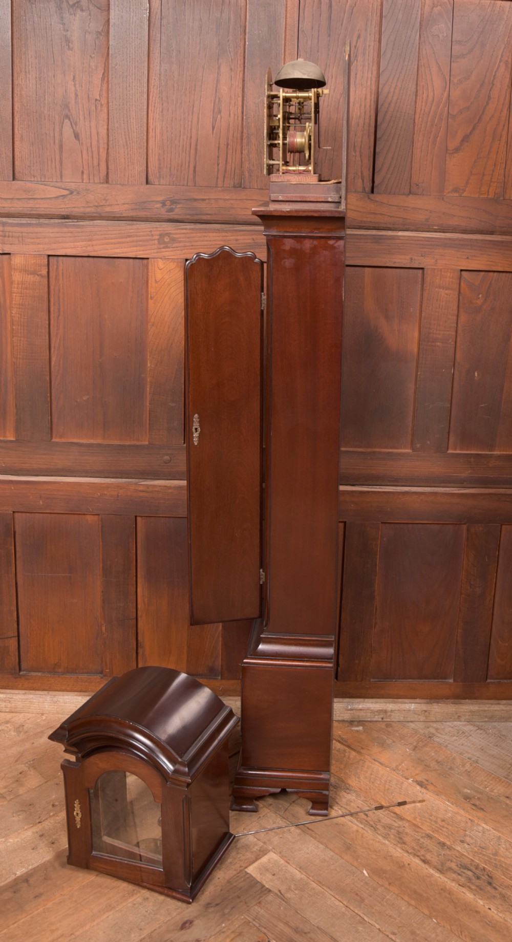 A freestanding mahogany grandmother clock featuring an arched hood, brass dial with corner spandrels, a slender waist door, and brass weights placed in front. View XI