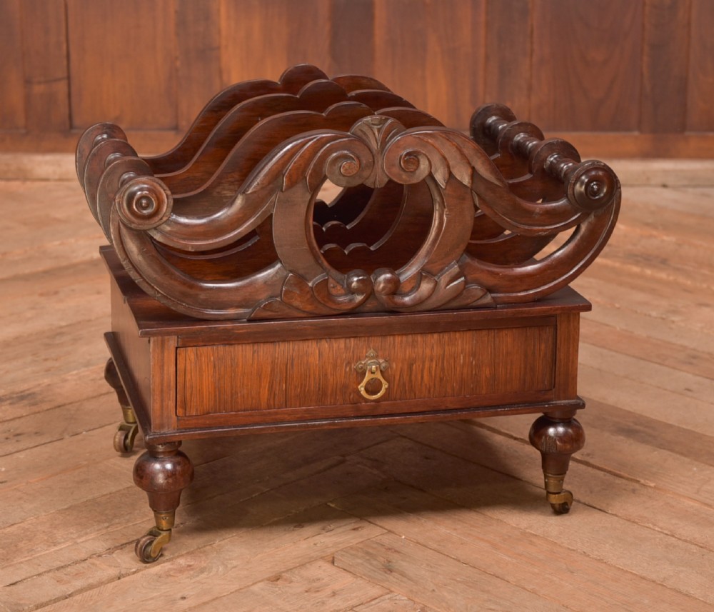 A Victorian rosewood Canterbury featuring four carved scroll dividers above a single base drawer, supported by turned legs on brass castors.