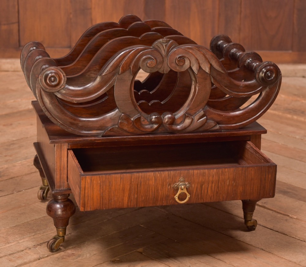 A Victorian rosewood Canterbury featuring four carved scroll dividers above a single base drawer, supported by turned legs on brass castors. View II