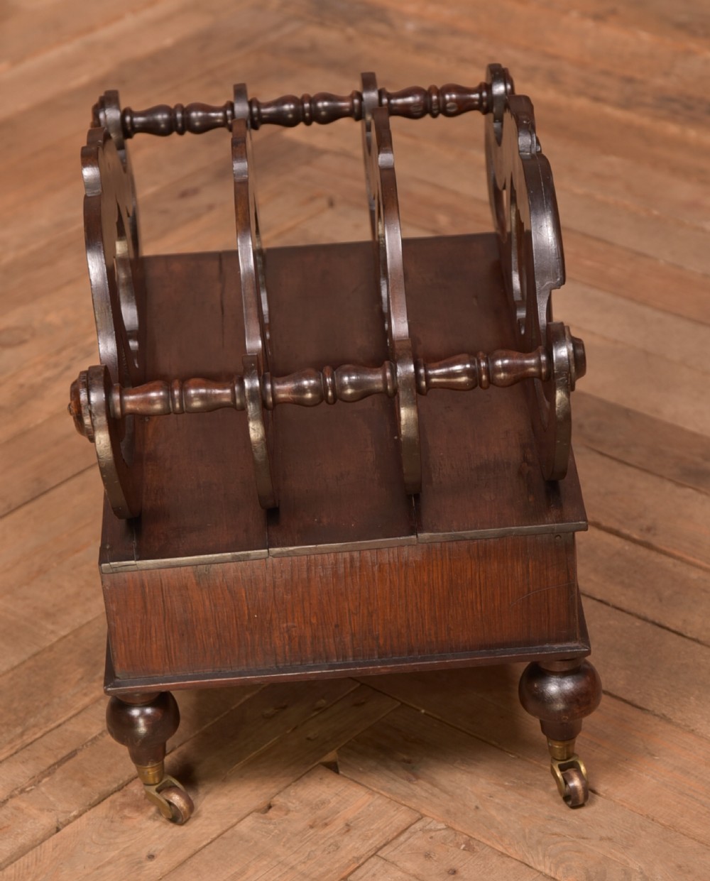 A Victorian rosewood Canterbury featuring four carved scroll dividers above a single base drawer, supported by turned legs on brass castors. View V