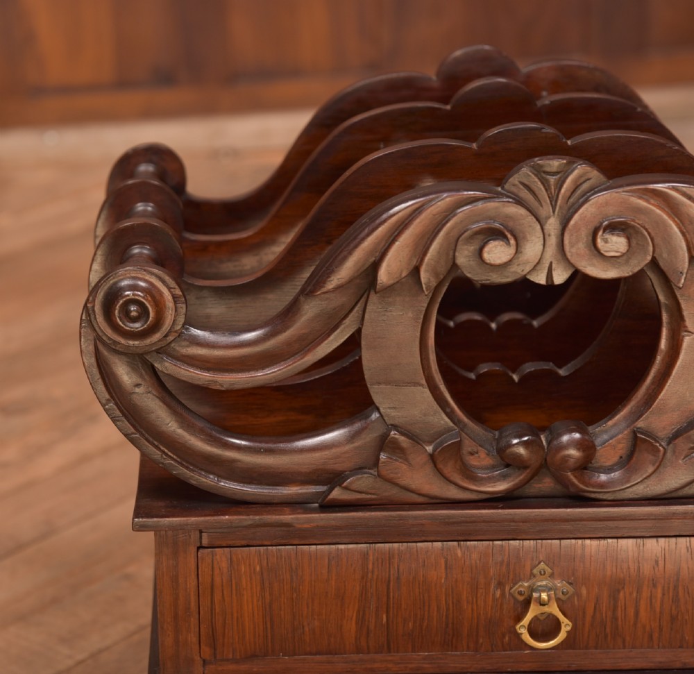 A Victorian rosewood Canterbury featuring four carved scroll dividers above a single base drawer, supported by turned legs on brass castors. View VI
