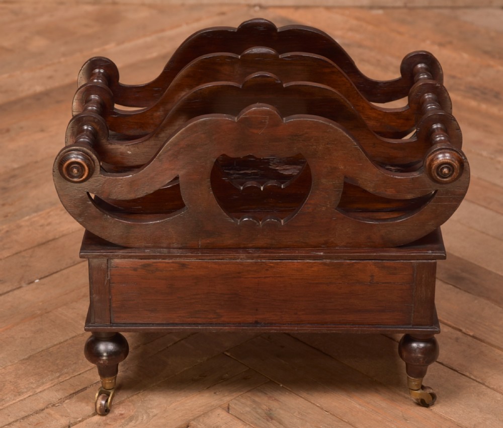 A Victorian rosewood Canterbury featuring four carved scroll dividers above a single base drawer, supported by turned legs on brass castors. View VII