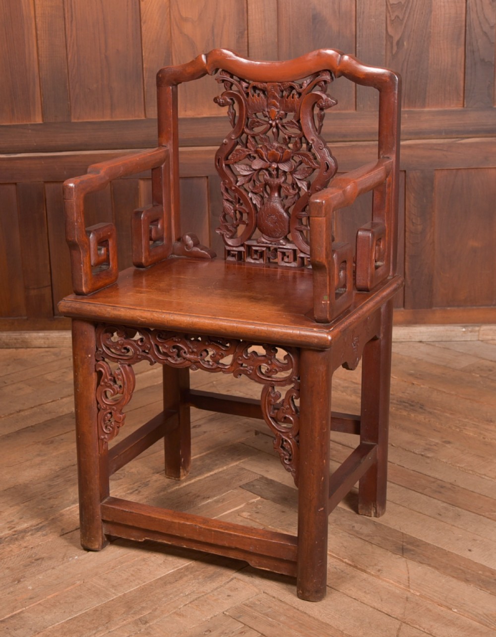 A carved reddish-brown lacquered wooden Chinese armchair featuring an intricate floral splat back, stepped armrests, and decorative carved apron.