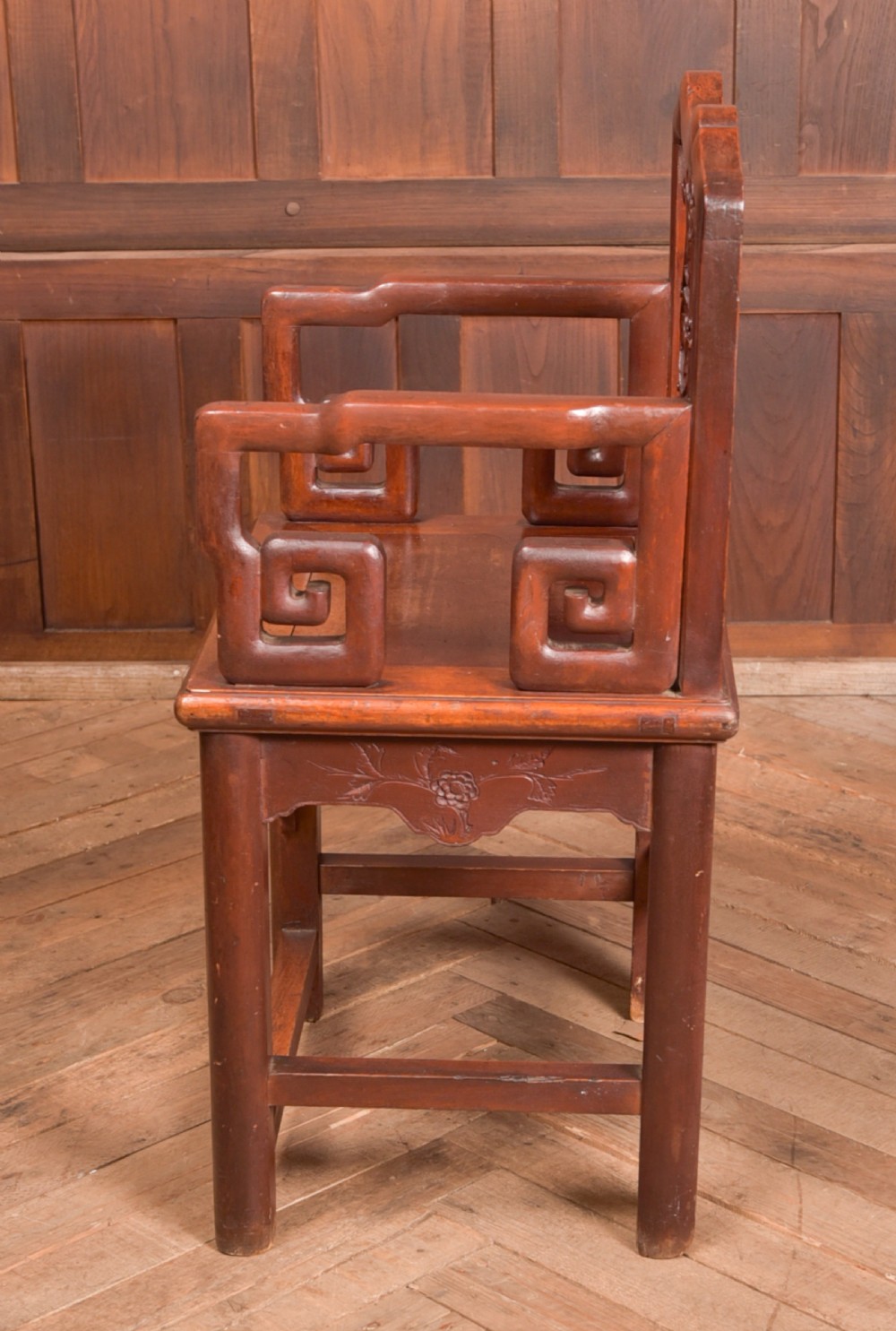 A carved reddish-brown lacquered wooden Chinese armchair featuring an intricate floral splat back, stepped armrests, and decorative carved apron. View V