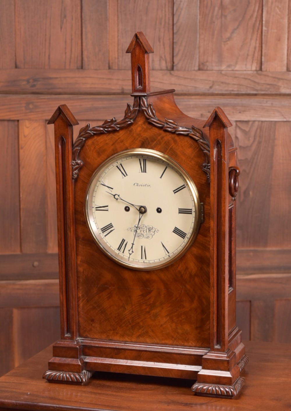 An antique mahogany bracket clock featuring Gothic spire finials, a carved foliage crest, and a cream dial signed Christie Perth set within a brass bezel.