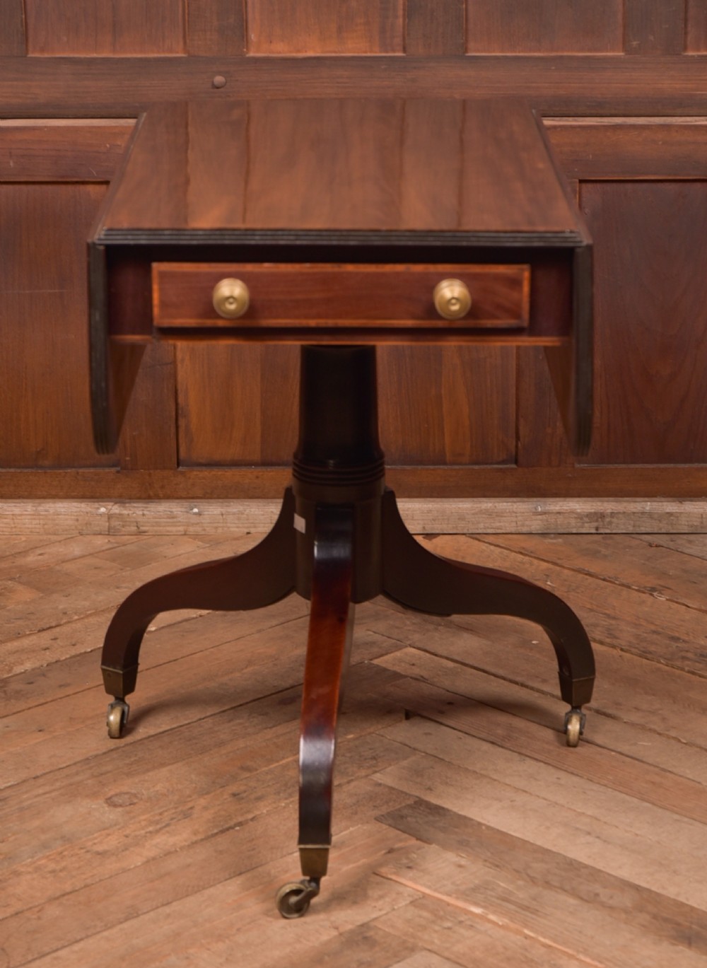 A dark mahogany Regency Pembroke table featuring a drop-leaf top, a single drawer with brass knobs, and a central turned pedestal resting on four splayed legs with brass castors. View III