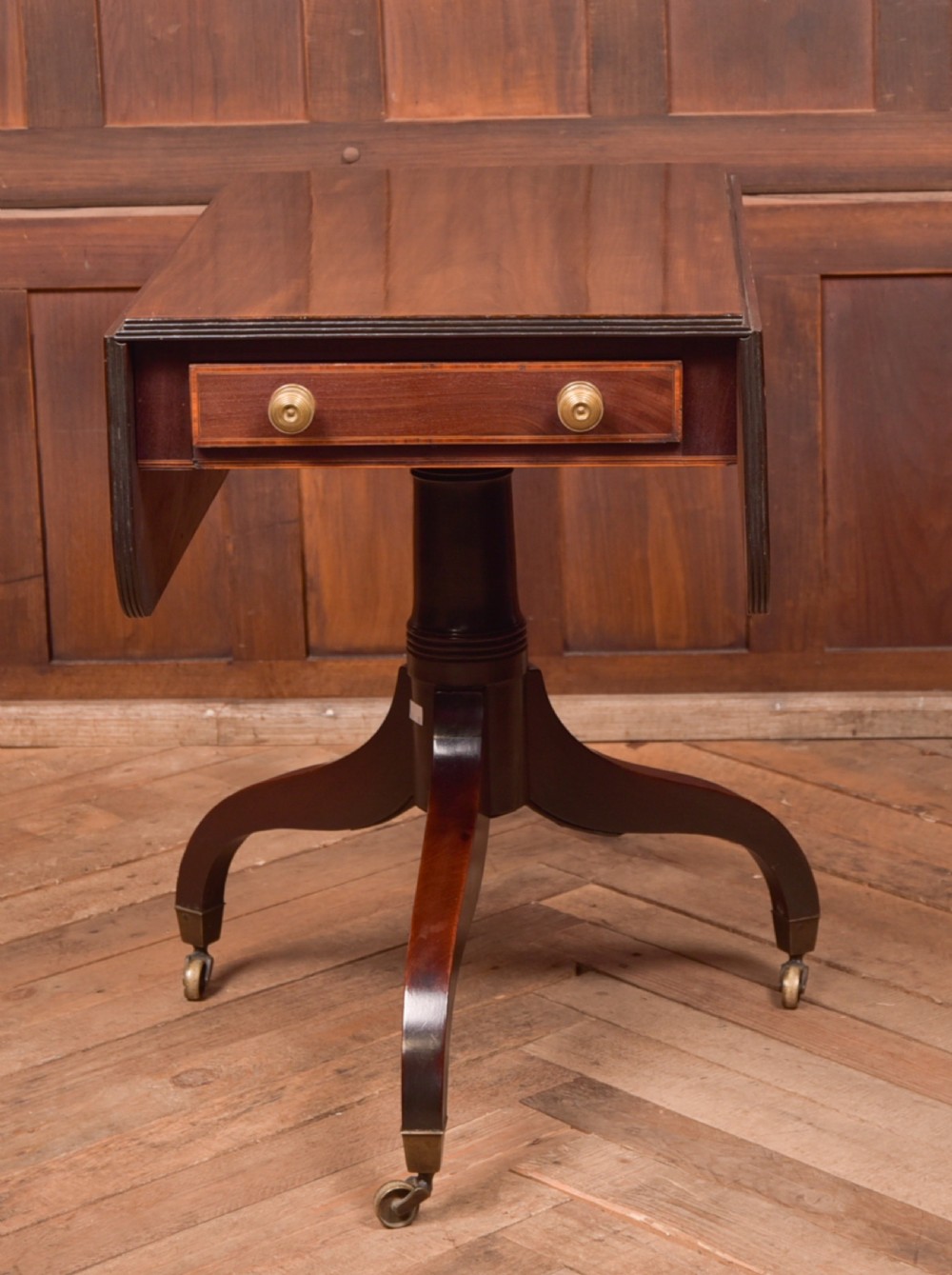 A dark mahogany Regency Pembroke table featuring a drop-leaf top, a single drawer with brass knobs, and a central turned pedestal resting on four splayed legs with brass castors. View IV