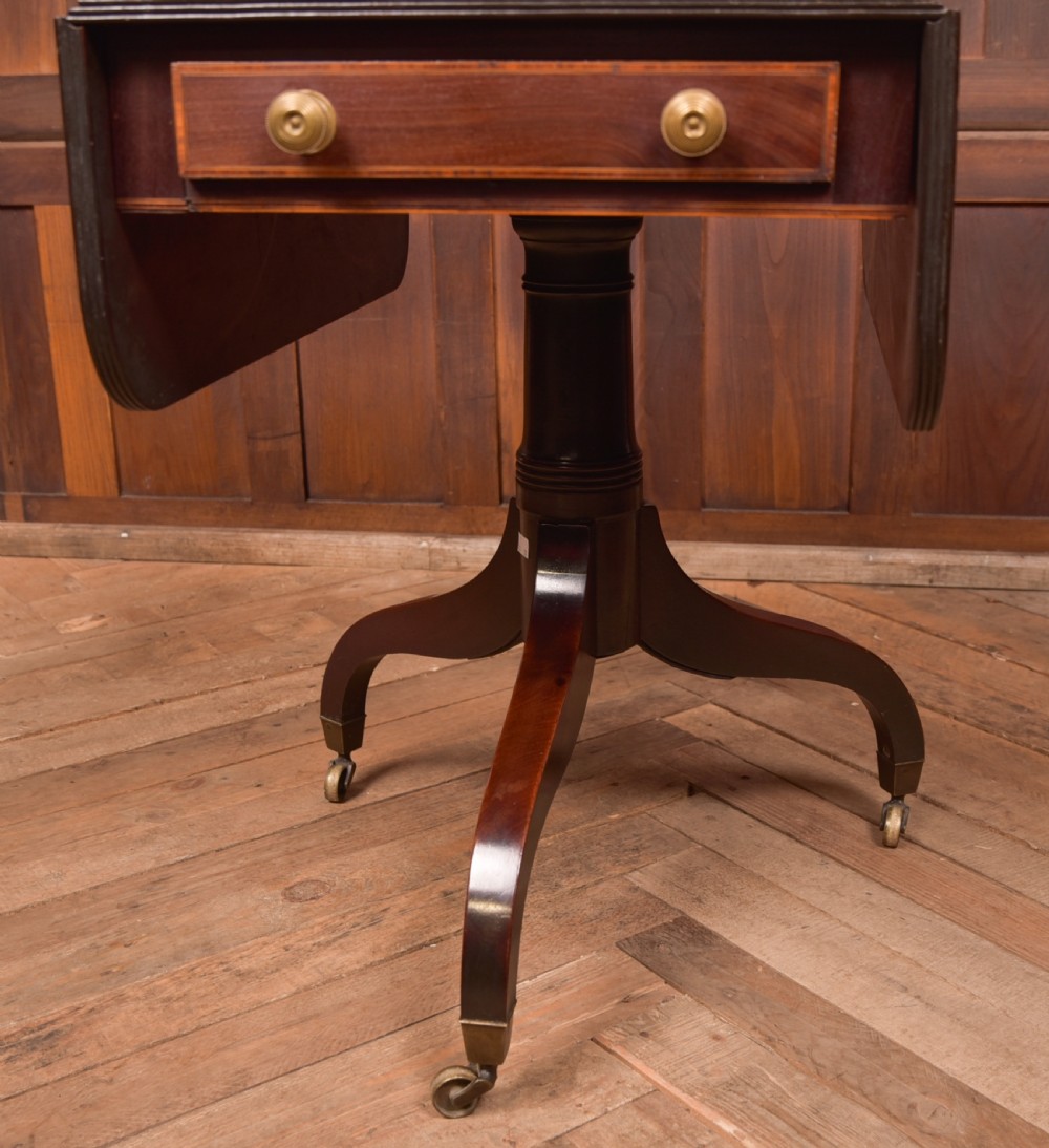 A dark mahogany Regency Pembroke table featuring a drop-leaf top, a single drawer with brass knobs, and a central turned pedestal resting on four splayed legs with brass castors. View VI