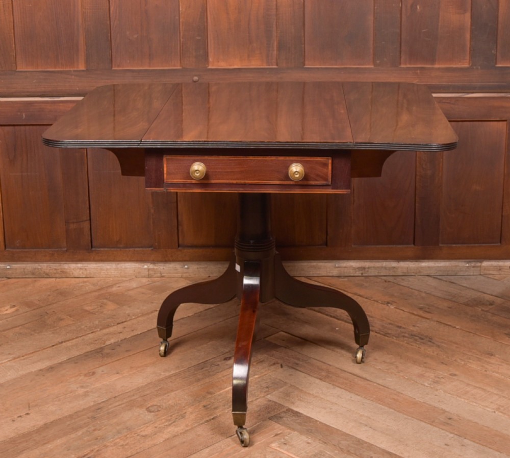 A dark mahogany Regency Pembroke table featuring a drop-leaf top, a single drawer with brass knobs, and a central turned pedestal resting on four splayed legs with brass castors. View VII