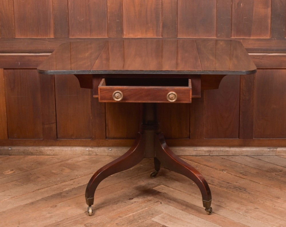 A dark mahogany Regency Pembroke table featuring a drop-leaf top, a single drawer with brass knobs, and a central turned pedestal resting on four splayed legs with brass castors. View IX