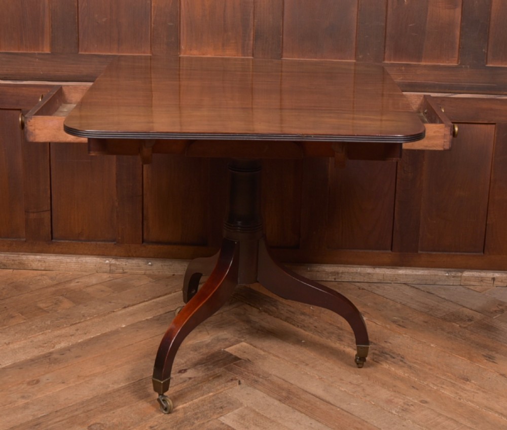 A dark mahogany Regency Pembroke table featuring a drop-leaf top, a single drawer with brass knobs, and a central turned pedestal resting on four splayed legs with brass castors. View X
