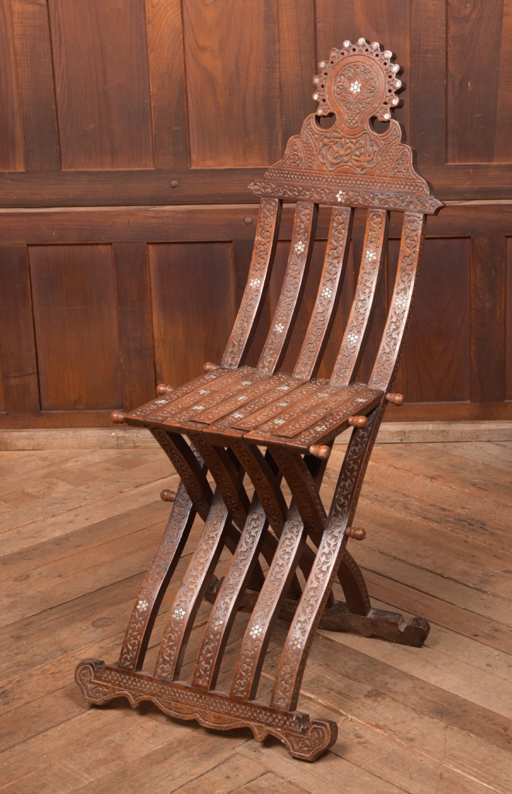 An intricate carved brown wooden Islamic folding chair decorated with mother-of-pearl inlay and calligraphic details.
