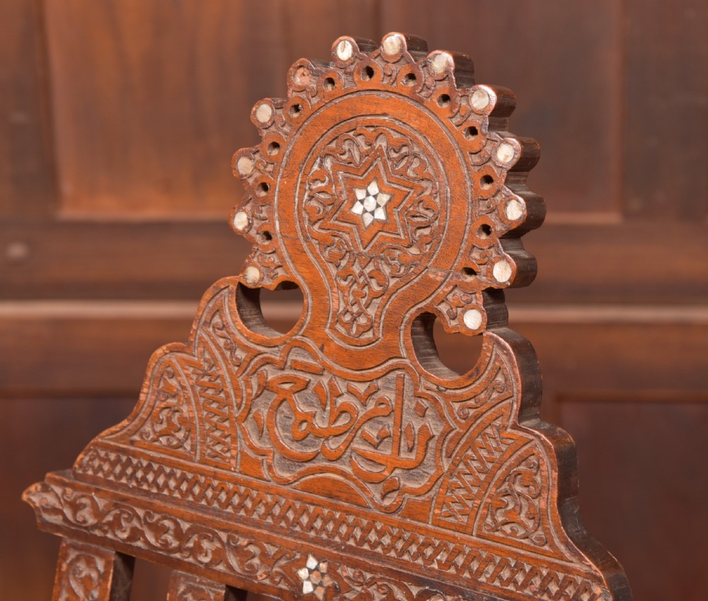 An intricate carved brown wooden Islamic folding chair decorated with mother-of-pearl inlay and calligraphic details. View II