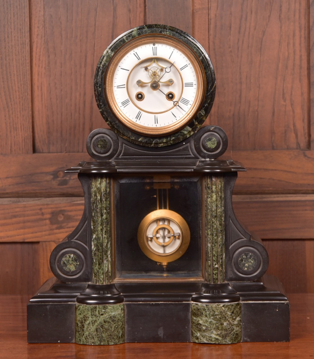 A Victorian French black slate and green marble mantel clock featuring a white enamel dial with Roman numerals and a visible brass pendulum behind glass.