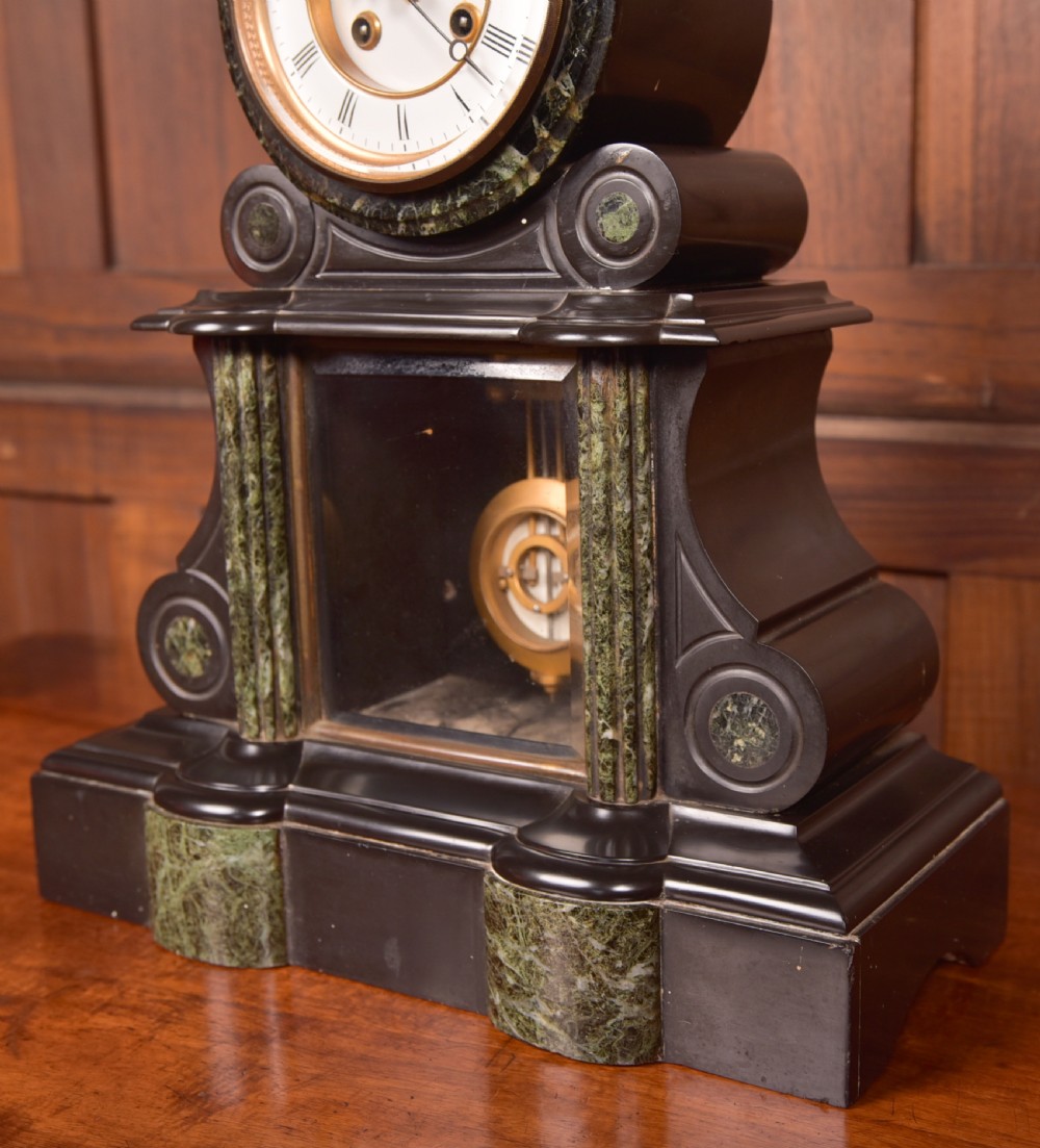 A Victorian French black slate and green marble mantel clock featuring a white enamel dial with Roman numerals and a visible brass pendulum behind glass. View II
