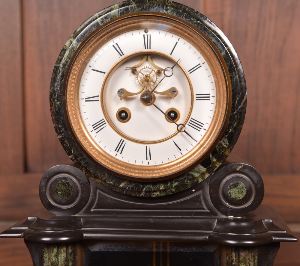 A Victorian French black slate and green marble mantel clock featuring a white enamel dial with Roman numerals and a visible brass pendulum behind glass. View V