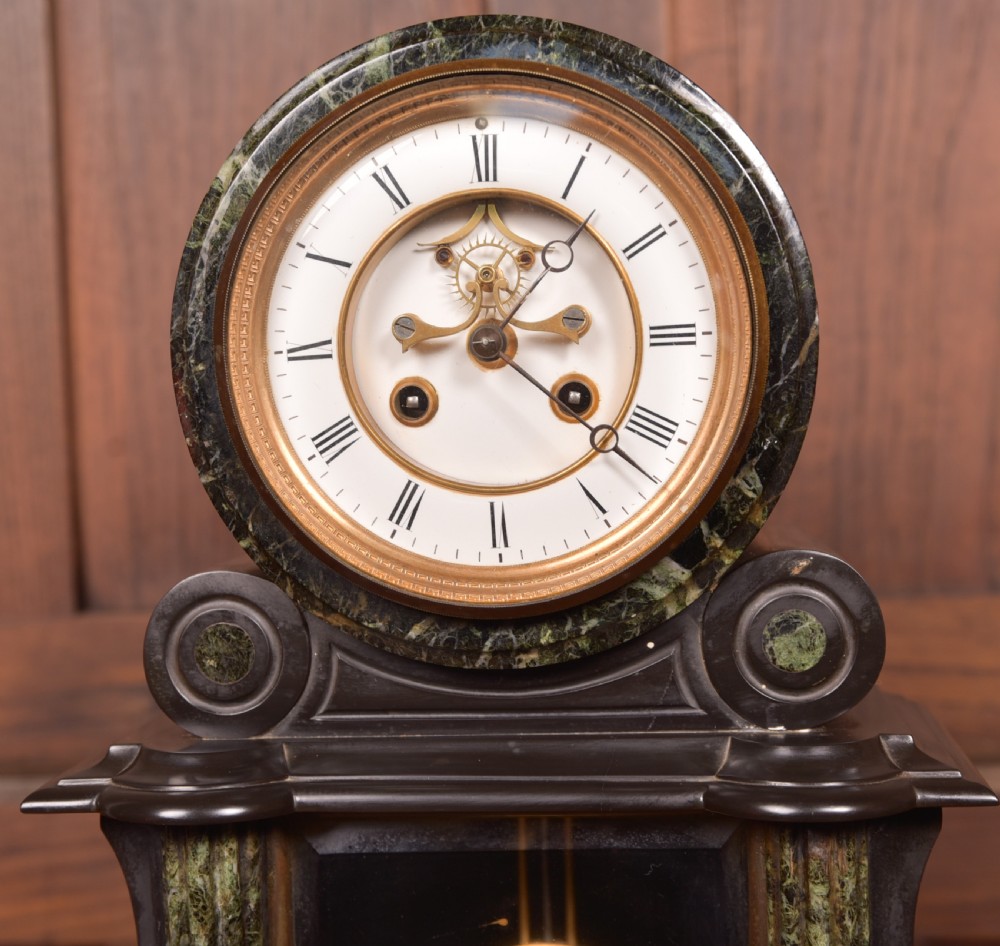 A Victorian French black slate and green marble mantel clock featuring a white enamel dial with Roman numerals and a visible brass pendulum behind glass. View VI