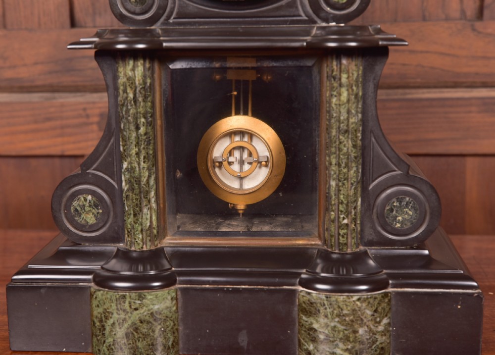 A Victorian French black slate and green marble mantel clock featuring a white enamel dial with Roman numerals and a visible brass pendulum behind glass. View VII