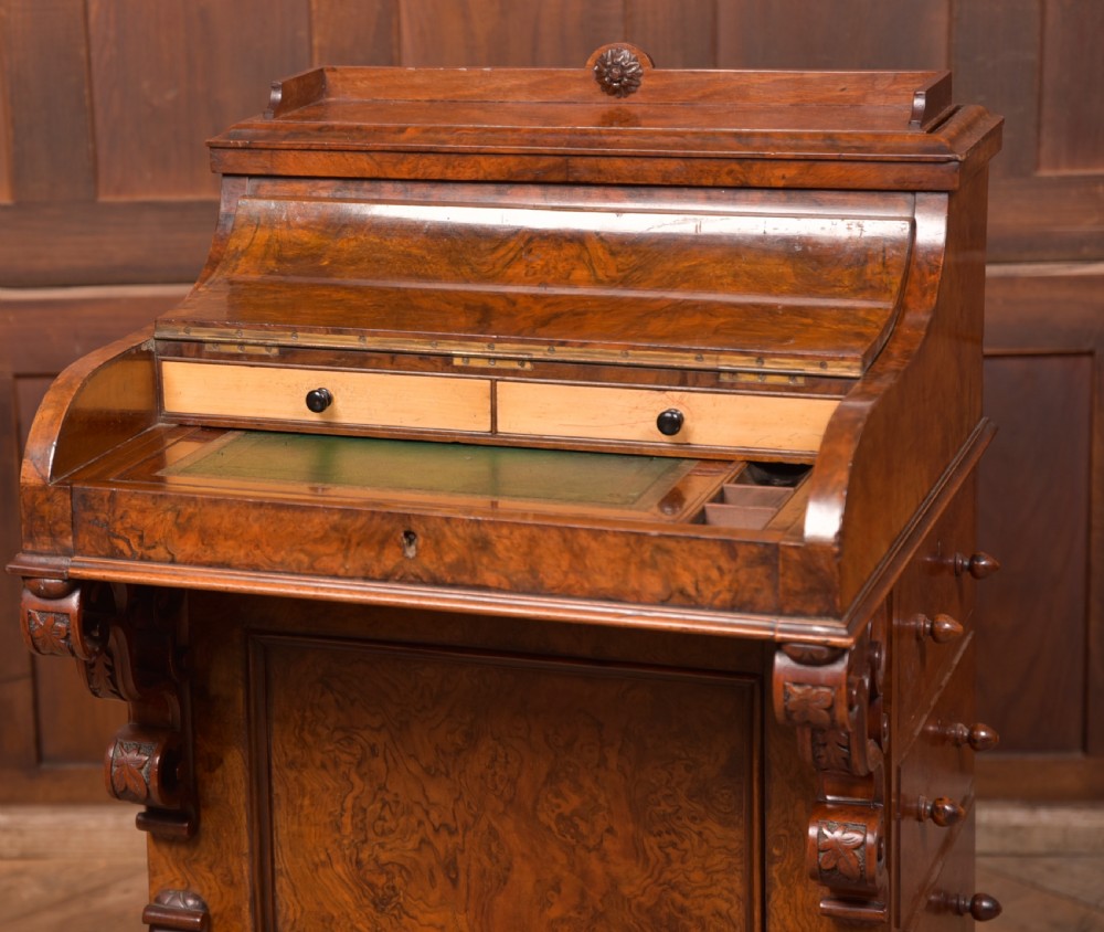 A Victorian burr walnut piano top davenport desk featuring a rounded sloping lid, carved scroll supports, and side drawers with turned wooden knobs. View V