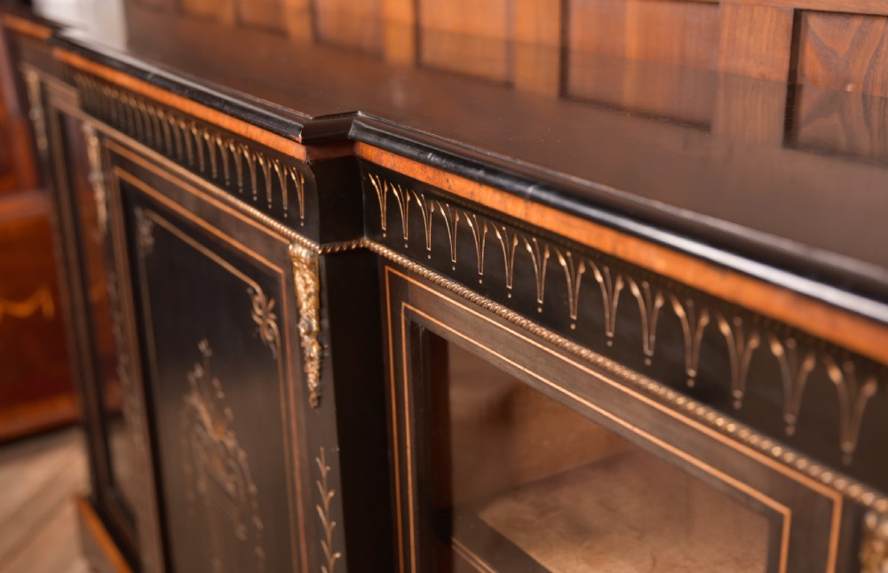 A Victorian ebonised breakfront credenza featuring glazed side doors, gilt incised urn motifs, and cast decorative mounts. View X