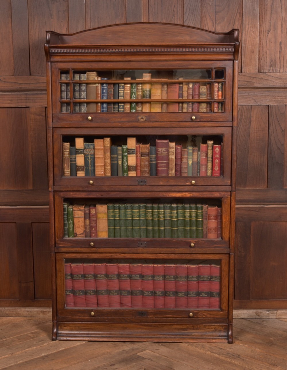 A four-tier dark oak sectional bookcase featuring glass front doors, brass hardware, and a decorative gallery top.