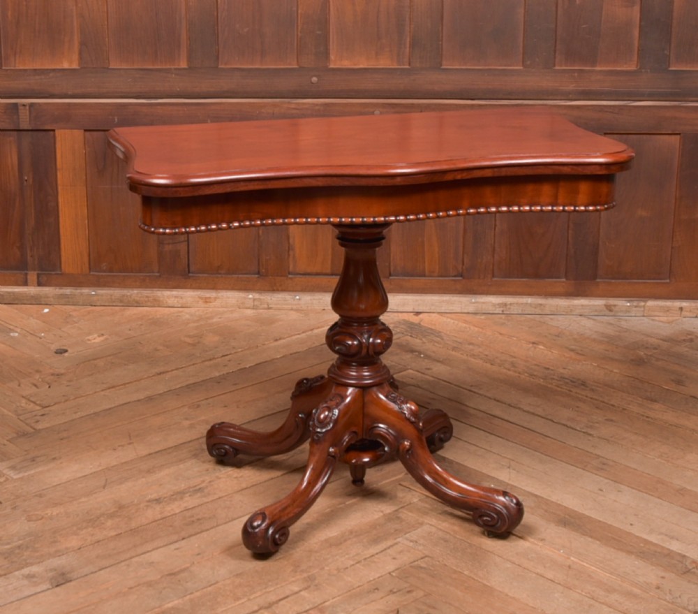 A Victorian mahogany fold-over tea table featuring a serpentine top, bead moulding along the apron, a turned central column, and carved quadruped scroll legs.