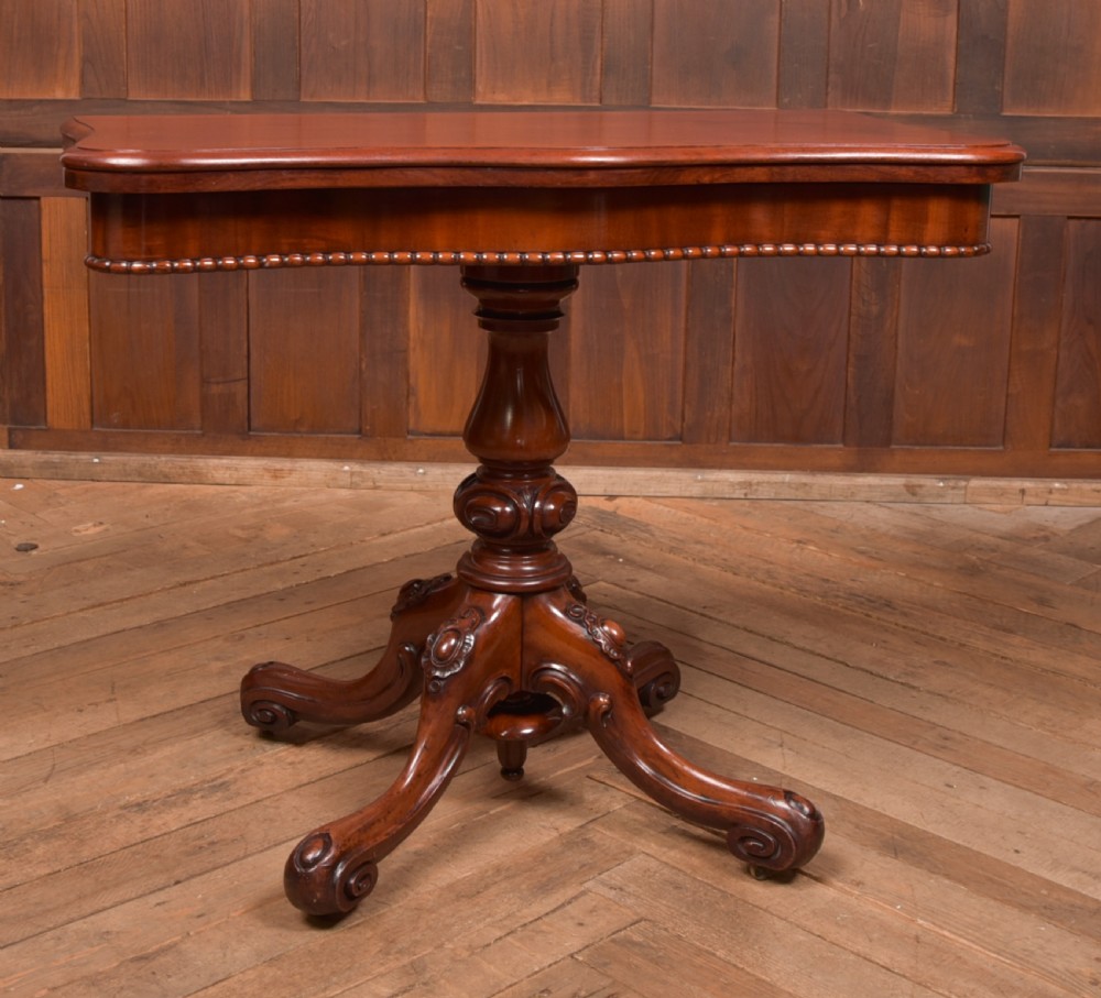 A Victorian mahogany fold-over tea table featuring a serpentine top, bead moulding along the apron, a turned central column, and carved quadruped scroll legs. View VII