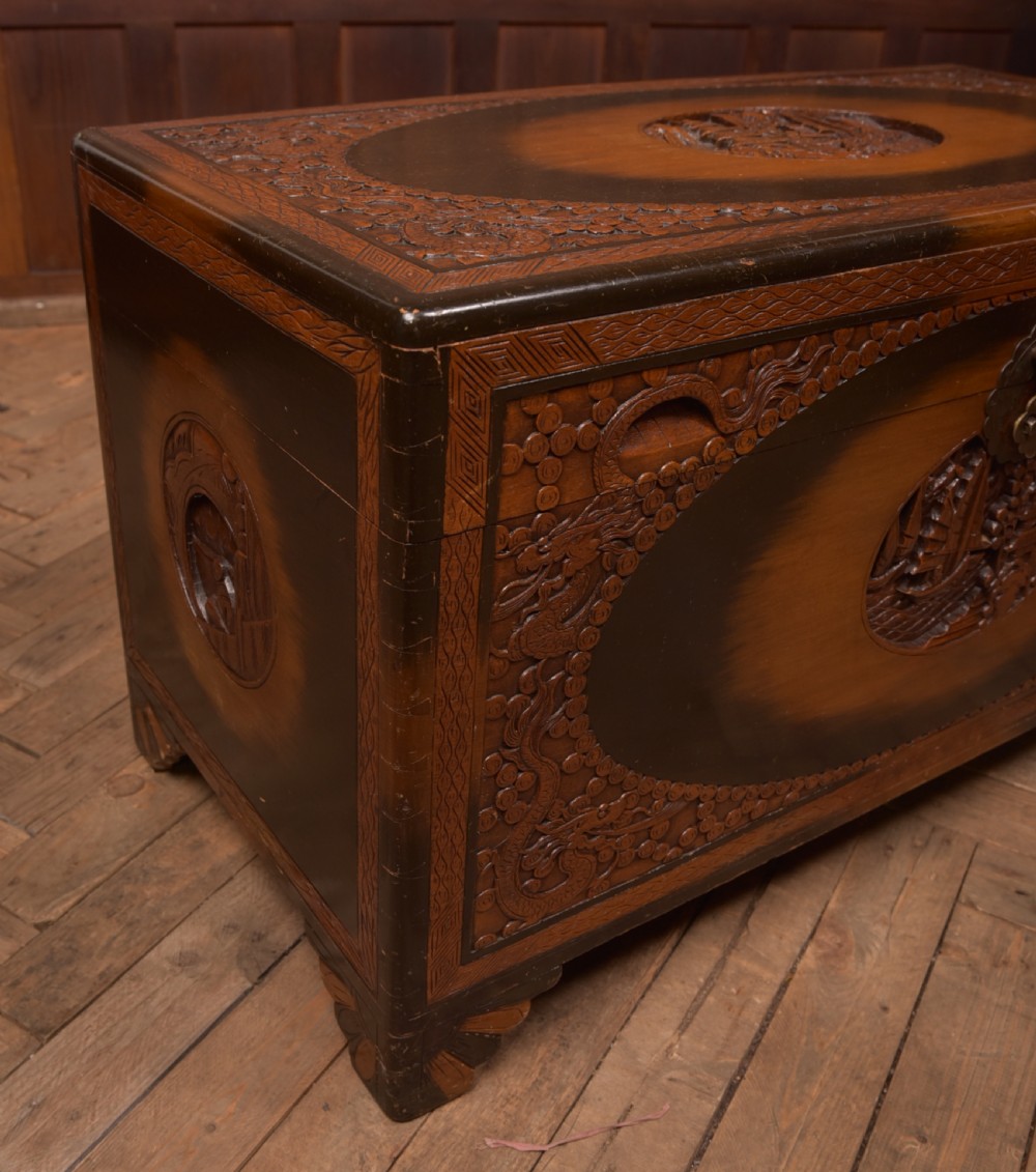 A hand-carved Chinese camphor wood storage chest featuring an oval relief medallion, dragon carvings, and a brass latch plate. View V