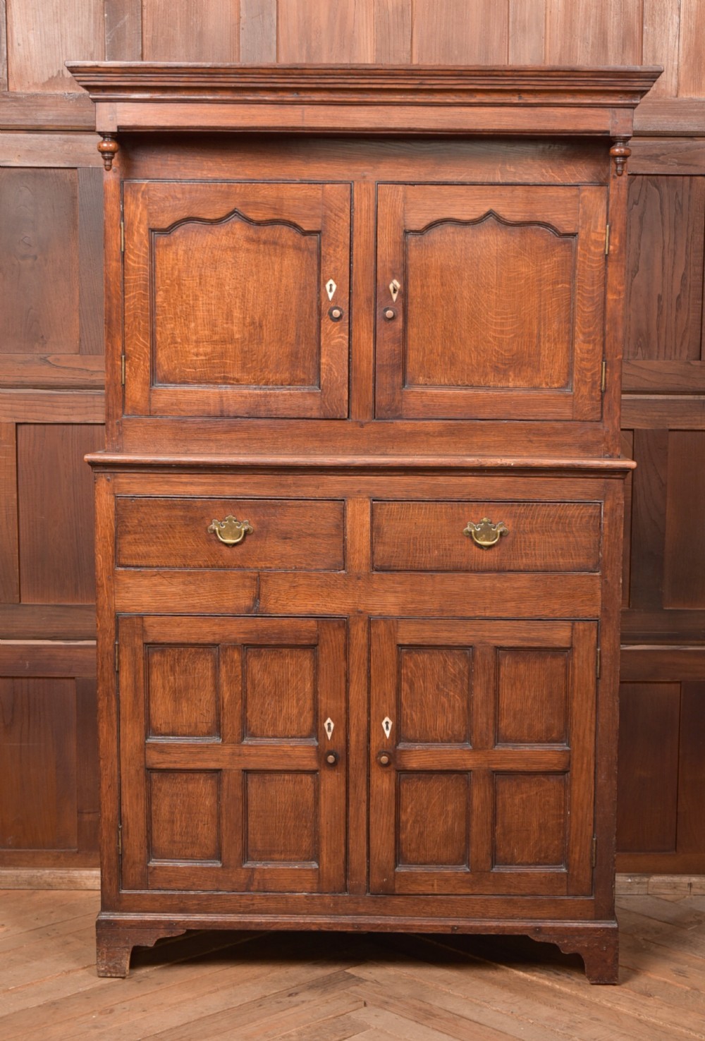 A two-tiered nineteenth-century oak court cupboard featuring panelled doors, drop handles on two upper drawers, and turned finials.
