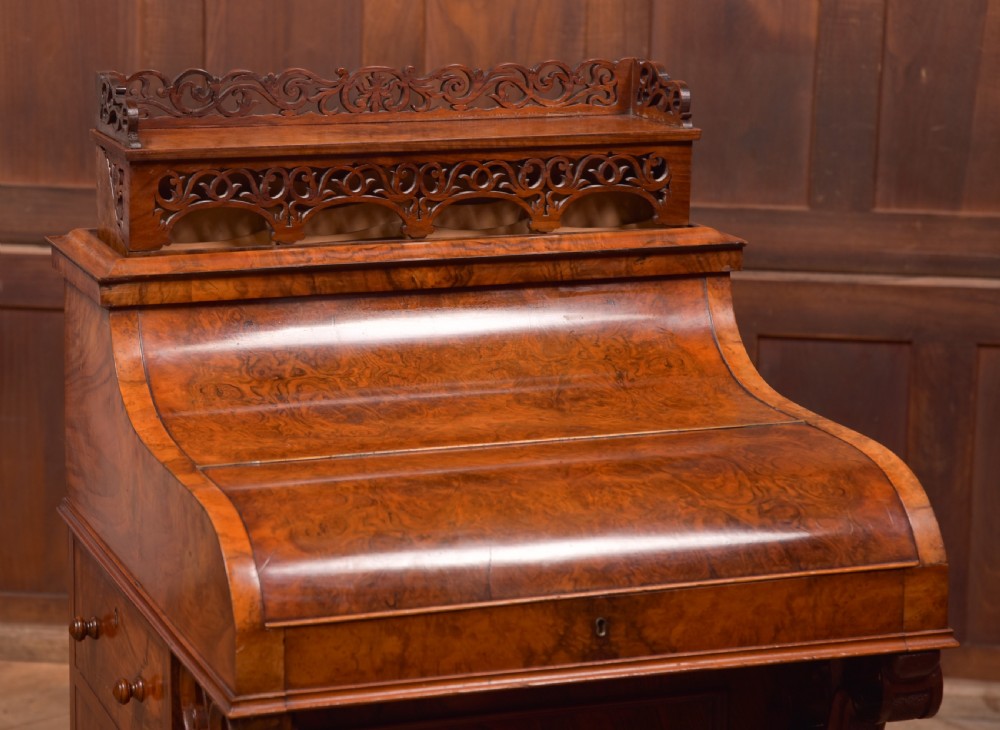 A Victorian burr walnut piano top Davenport desk featuring a carved fretwork gallery, sloped writing lid, scroll front supports, and side-opening drawers. View VI