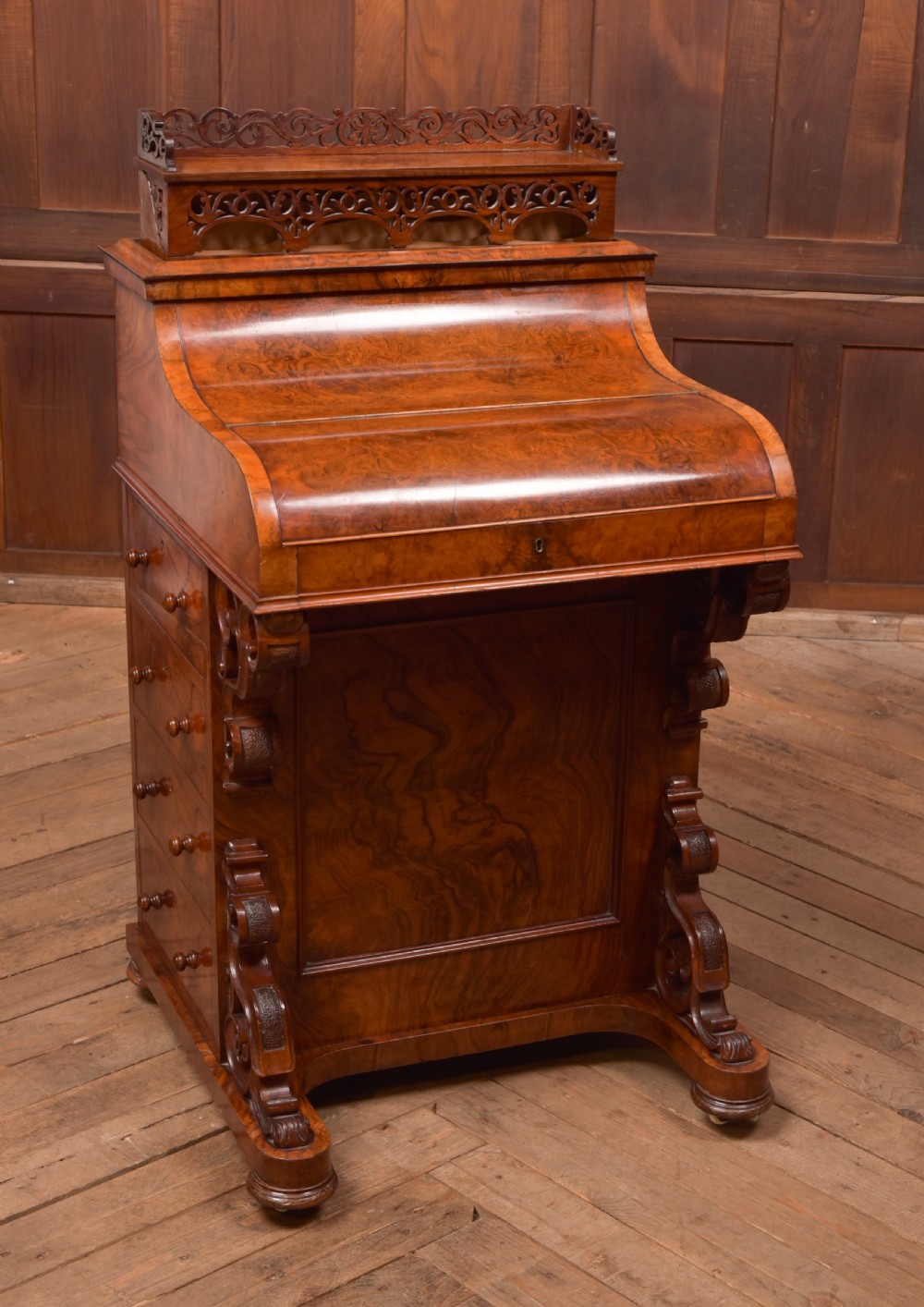 A Victorian burr walnut piano top Davenport desk featuring a carved fretwork gallery, sloped writing lid, scroll front supports, and side-opening drawers. View XVI