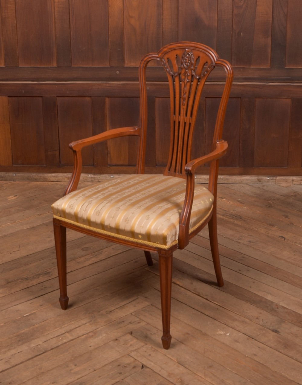An Edwardian satinwood armchair with a carved pierced splat back, sweeping armrests, and a striped gold upholstered seat.