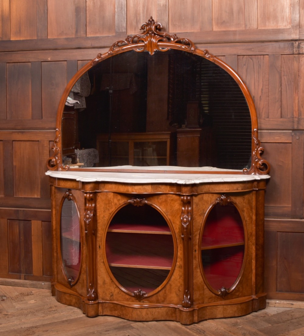 A Victorian burr walnut mirror-back credenza featuring a shaped white marble top, carved scrollwork crest, and three oval glazed cabinet doors revealing red fabric lining.