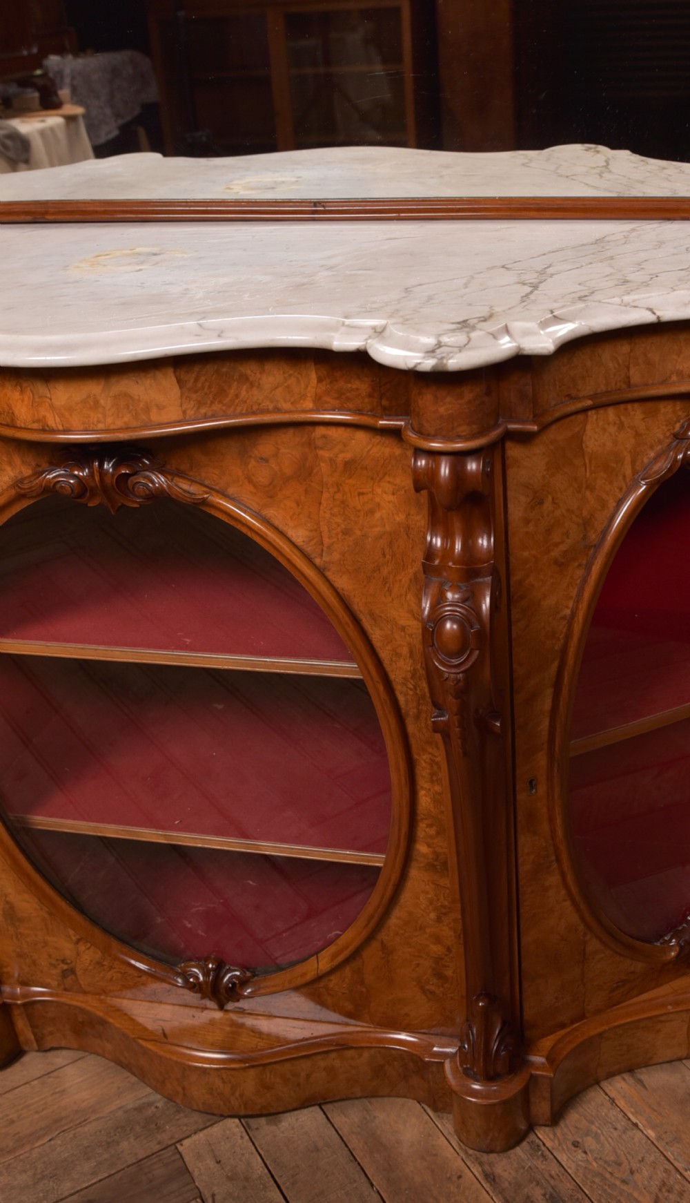 A Victorian burr walnut mirror-back credenza featuring a shaped white marble top, carved scrollwork crest, and three oval glazed cabinet doors revealing red fabric lining. View VI