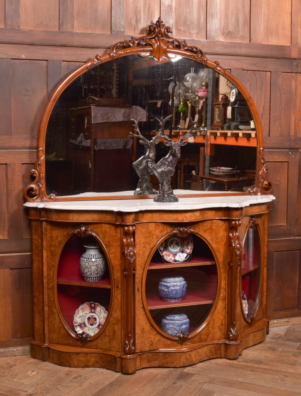 A Victorian burr walnut mirror-back credenza featuring a shaped white marble top, carved scrollwork crest, and three oval glazed cabinet doors revealing red fabric lining. View XV