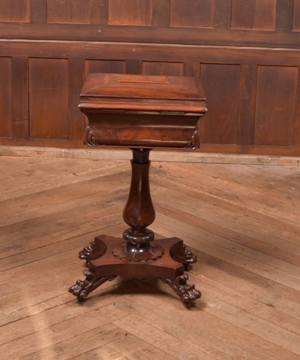 A Victorian rosewood work table featuring a hinged sarcophagus lid, a turned pedestal column, and a carved quadruped base on brass castors. View X