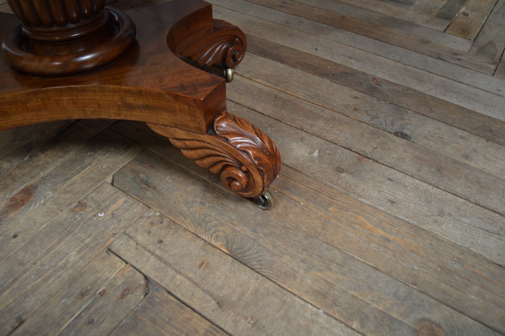 A William IV mahogany fold-over tea table featuring a swivelling top, a fluted turned central pedestal, and a quadform base with scroll feet on brass castors. View XIII