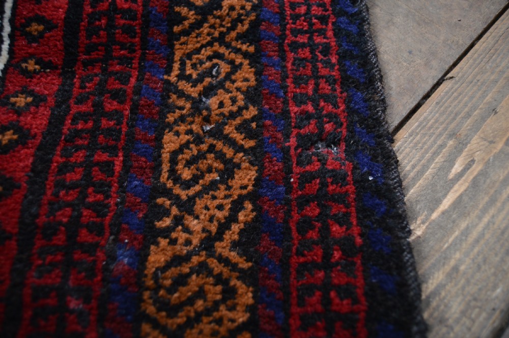 A hand-woven Belouch wool prayer rug featuring a central ochre mihrab motif surrounded by dark red and geometric patterned borders. View V