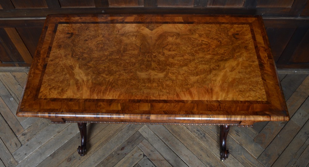 A Victorian burr walnut side table featuring a crossbanded rectangular top, two turned pedestals with a central stretcher, and carved legs with castors. View XV