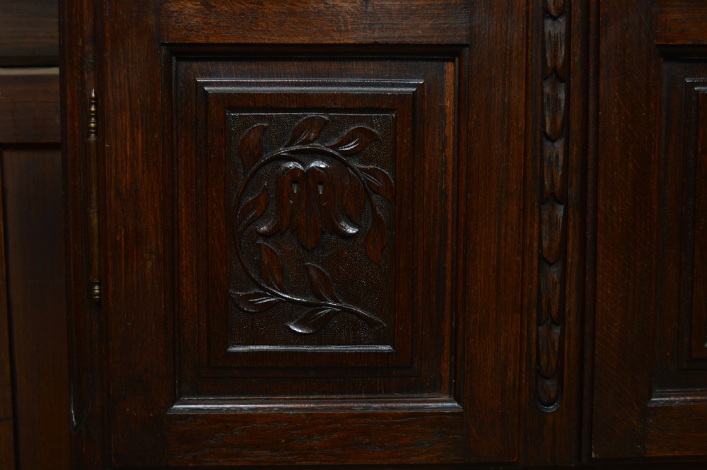 A dark carved oak display cabinet featuring two glazed doors with upper floral carvings and central brass key escutcheons. View V