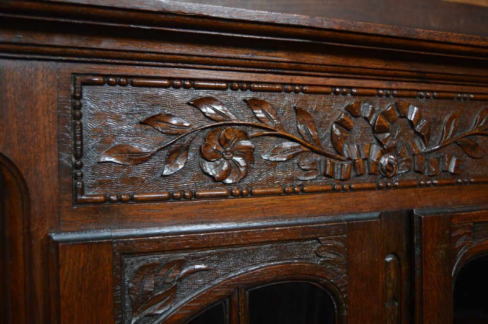 A dark carved oak display cabinet featuring two glazed doors with upper floral carvings and central brass key escutcheons. View VII