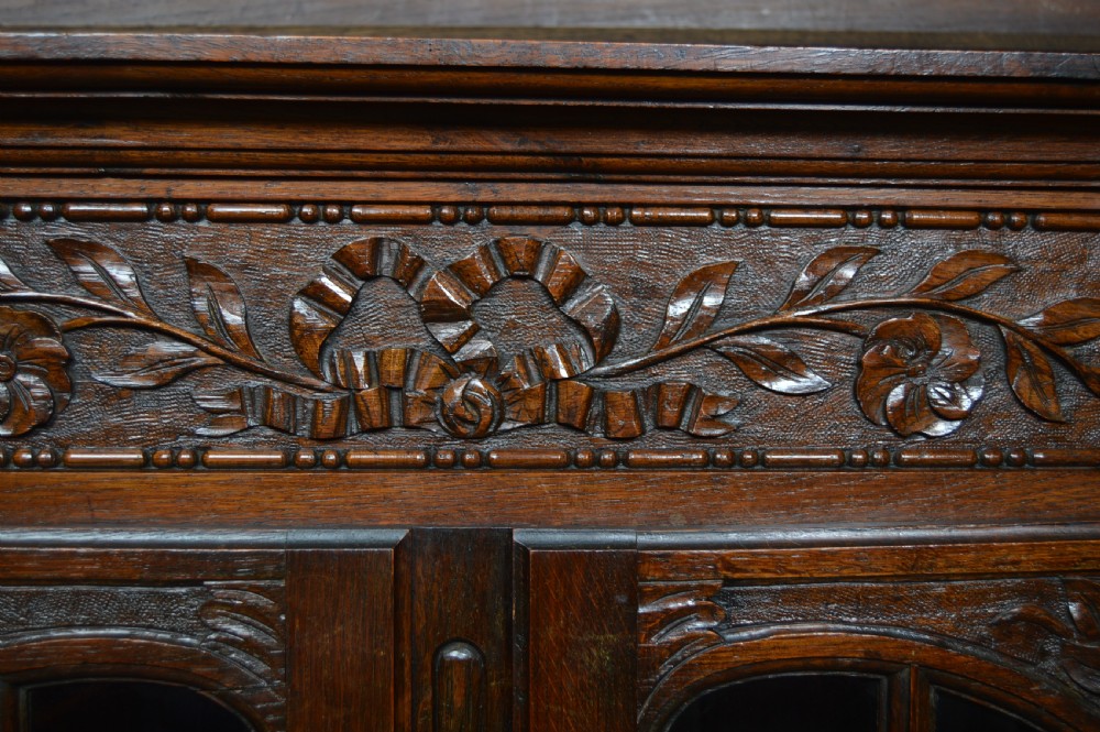 A dark carved oak display cabinet featuring two glazed doors with upper floral carvings and central brass key escutcheons. View VIII