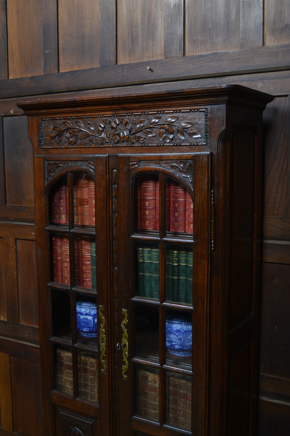 A dark carved oak display cabinet featuring two glazed doors with upper floral carvings and central brass key escutcheons. View XXI