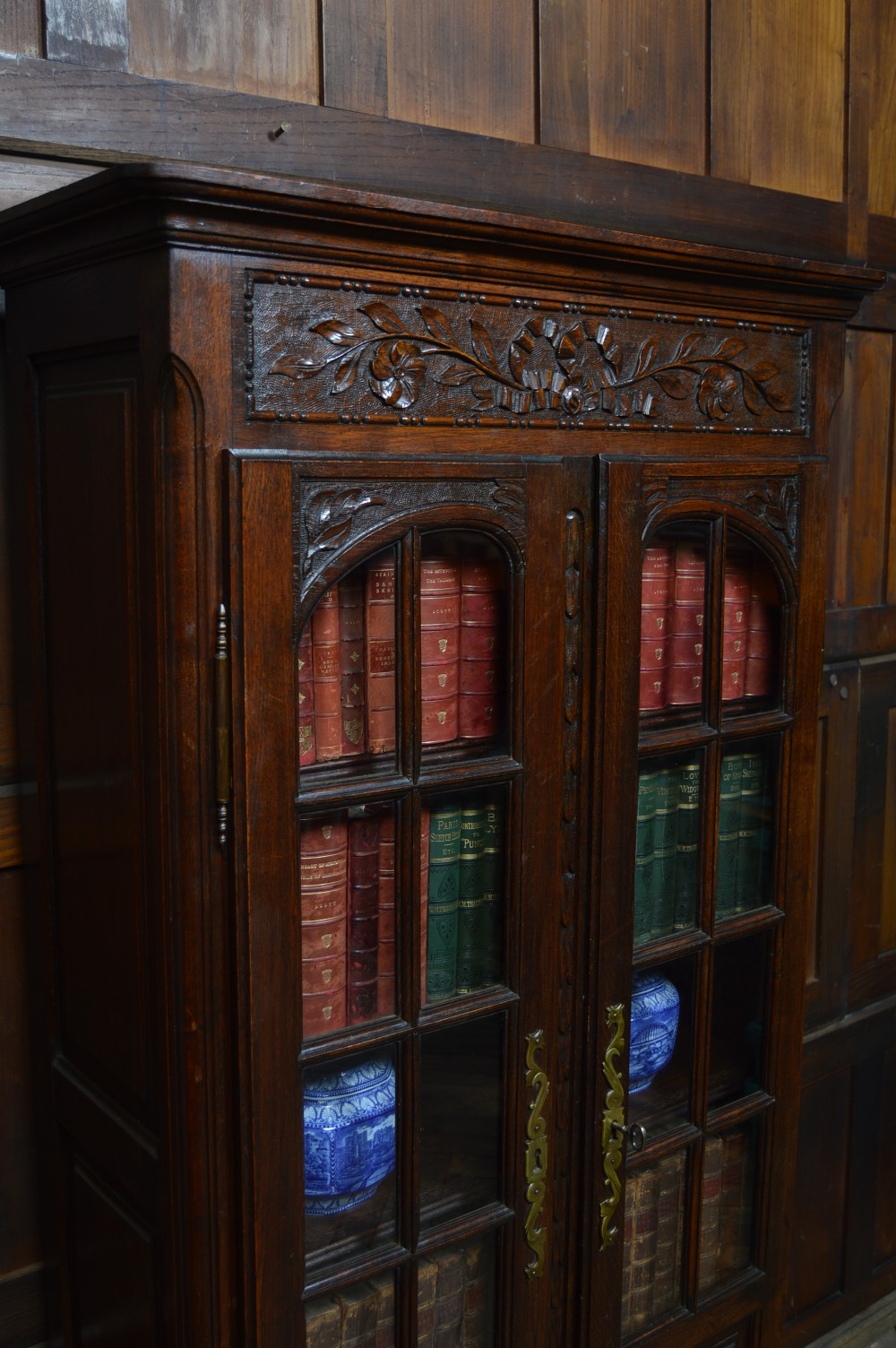 A dark carved oak display cabinet featuring two glazed doors with upper floral carvings and central brass key escutcheons. View XXII