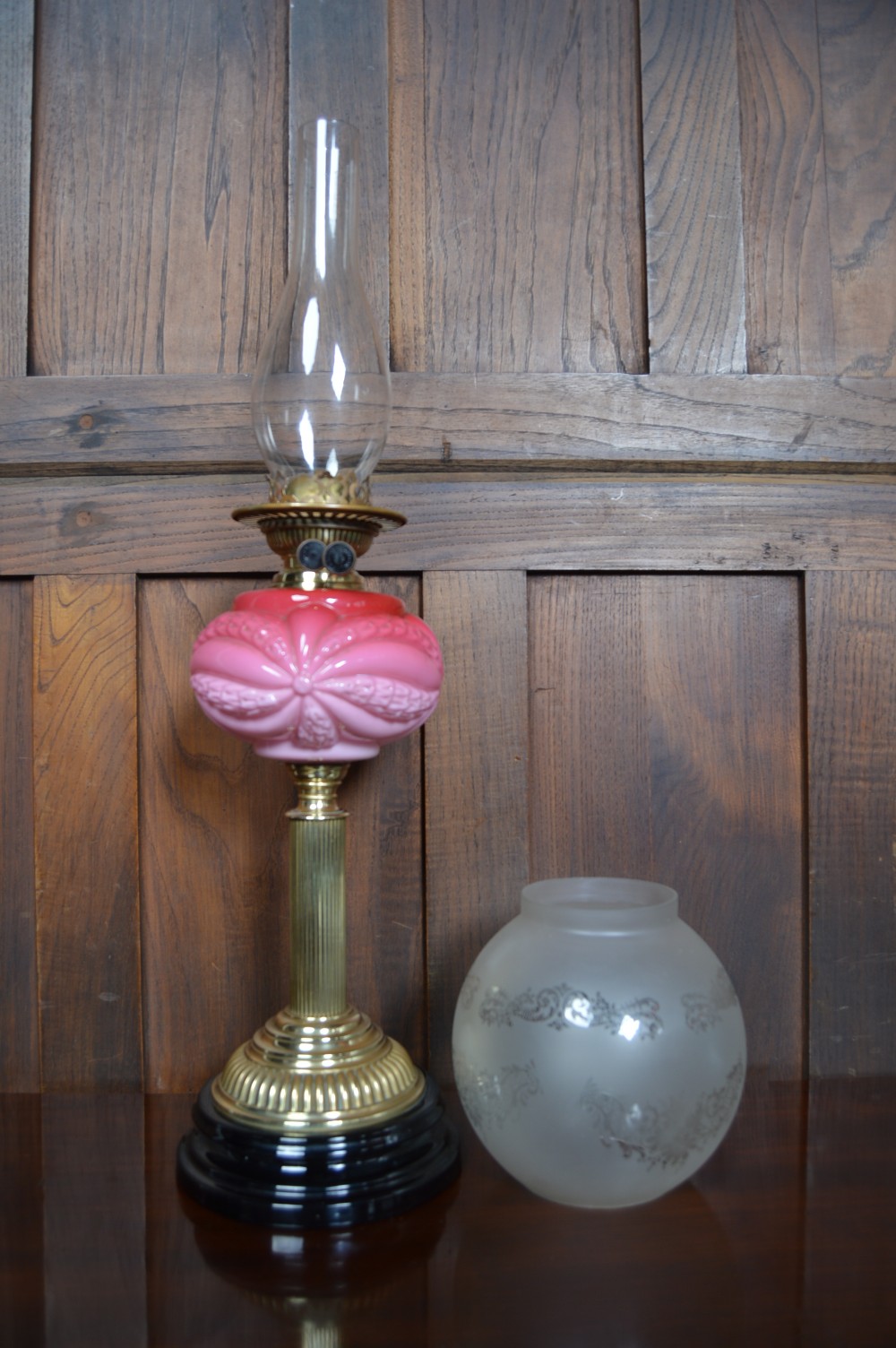 A Victorian brass oil lamp with a black base, fluted column, pink patterned glass reservoir, and frosted glass globe shade with etched detail. View VIII